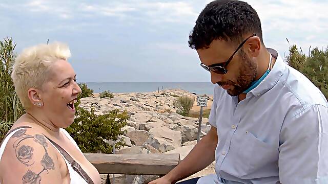 A busty blonde in a summer dress walks along the road, looking for a ride.